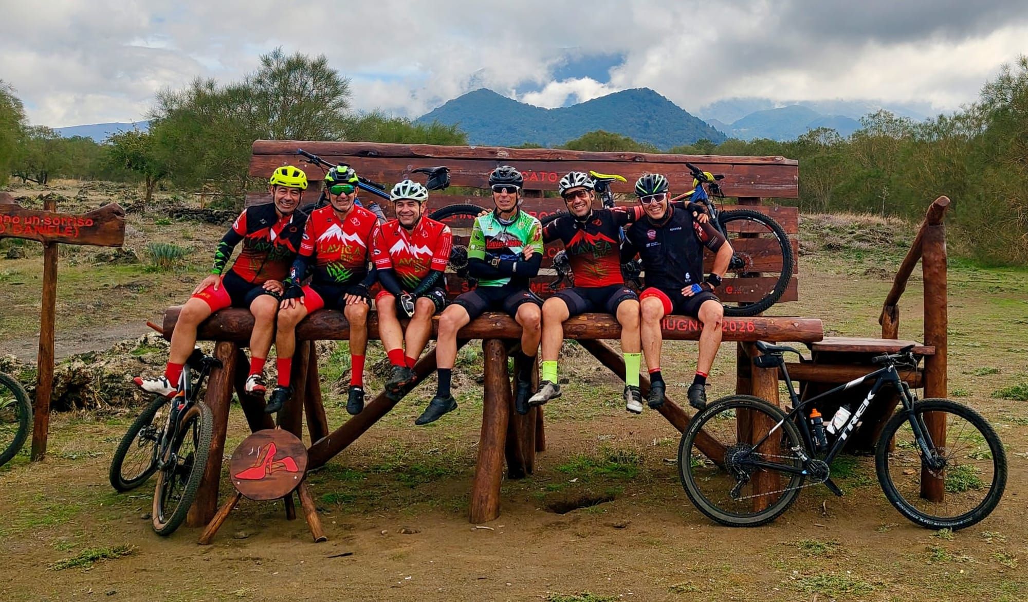 Sei ciclisti seduti su una panchina gigante di legno, con le bici appoggiate e i monti sullo sfondo