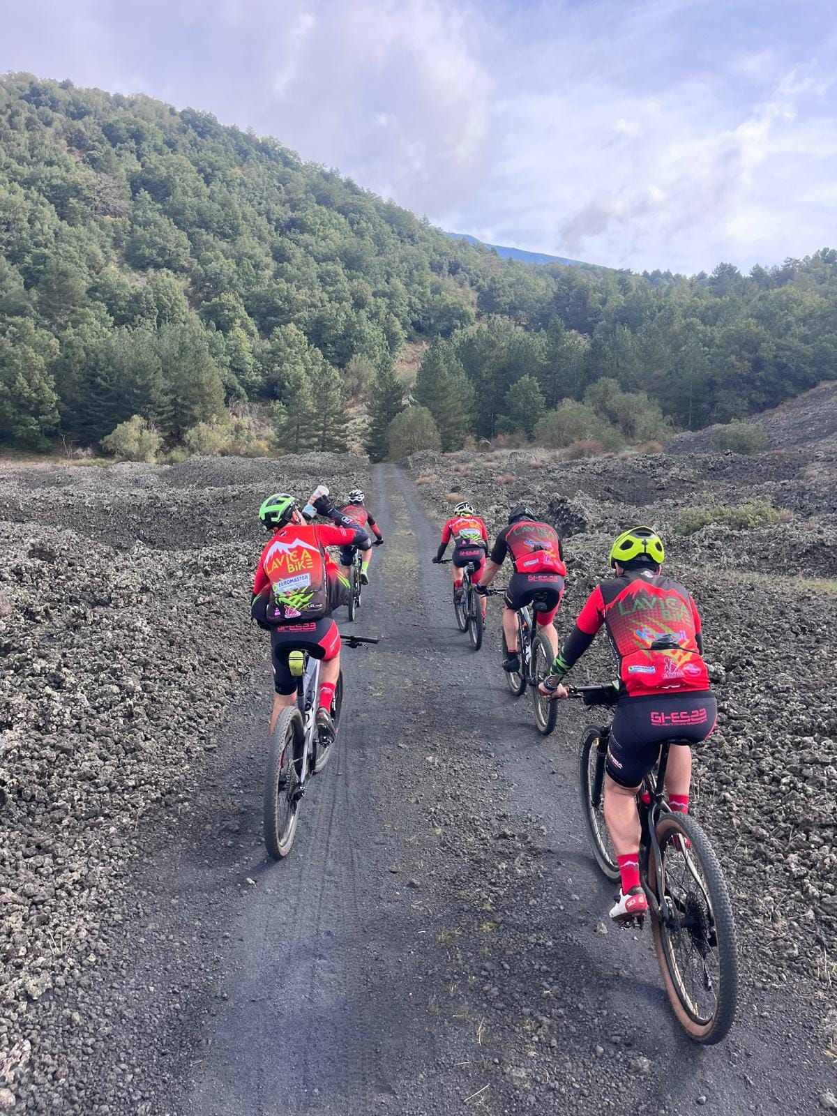 Cinque ciclisti in maglia rossa salgono su una pista sterrata tra la sabbia lavica, verso il bosco
